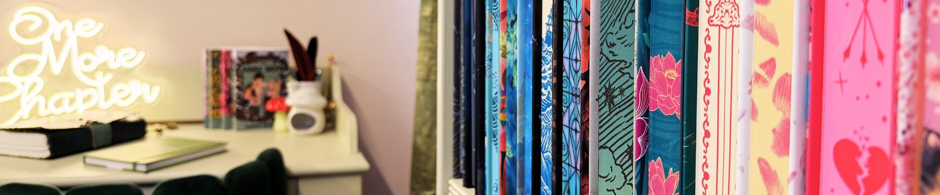 Reading nook ideas with a blush pink writing space, glowing “One More Chapter” sign, green velvet desk chair, and a bookshelf lined with colourful special edition books.