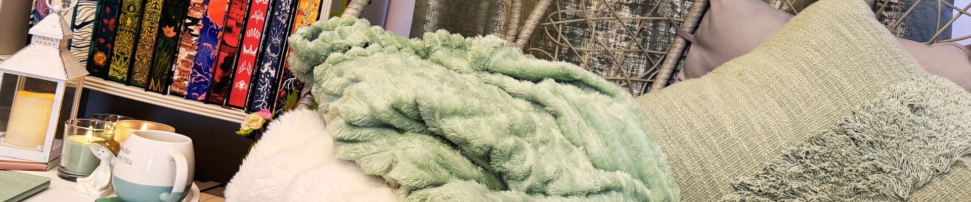 Cozy reading nook ideas with a hanging egg chair layered with sage green cushions and blankets, candlelight, a mug, lantern decor, and shelves of colourful special edition books.