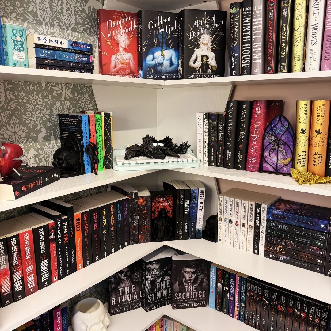 Cozy reading nook ideas in a book-filled corner with white corner shelves, sage patterned wallpaper, stacked fantasy and dark romance books, a purple stained-glass style book nook insert, skull decor, and black dragon display accents.