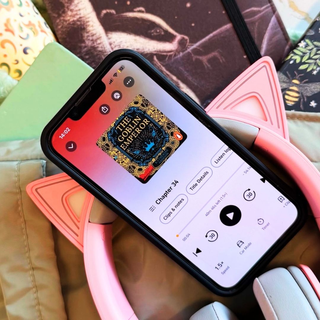 Phone playing The Goblin Emperor by Katherine Addison audiobook for a review, with pink headphones and notebooks nearby, including one with a badger on the cover.