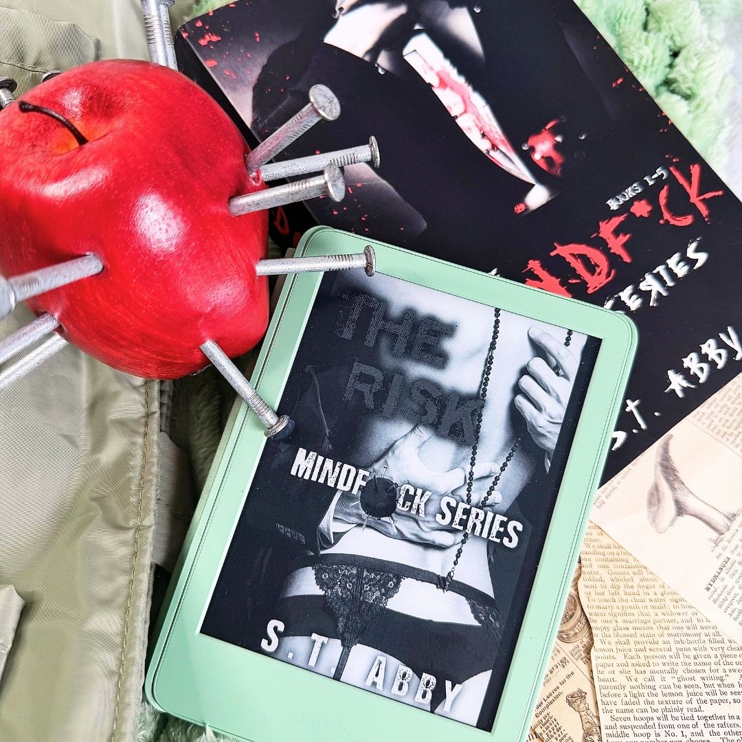 Flat lay of The Risk by S.T. Abby, Book 1 of the Mindfuck series. The Risk's cover is visible on a mint green Kindle, surrounded by dark romance props including a red apple pierced with nails, a bloody knife book cover, and torn book pages. The kindle rests on a paperback omnibus edition of the full Mindf*ck series.