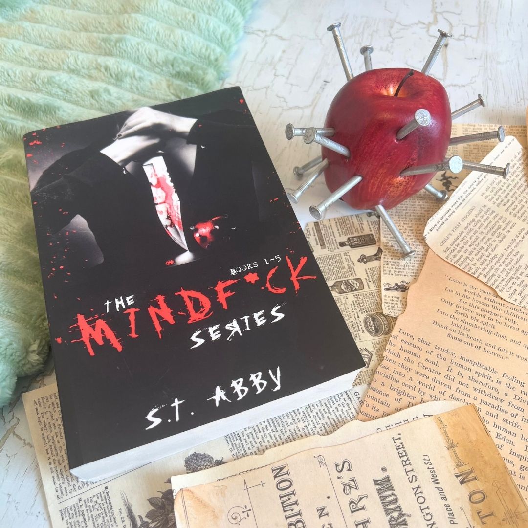 The Mindfuck Series by S.T. Abby beside a red apple pierced with nails on scattered vintage book pages.